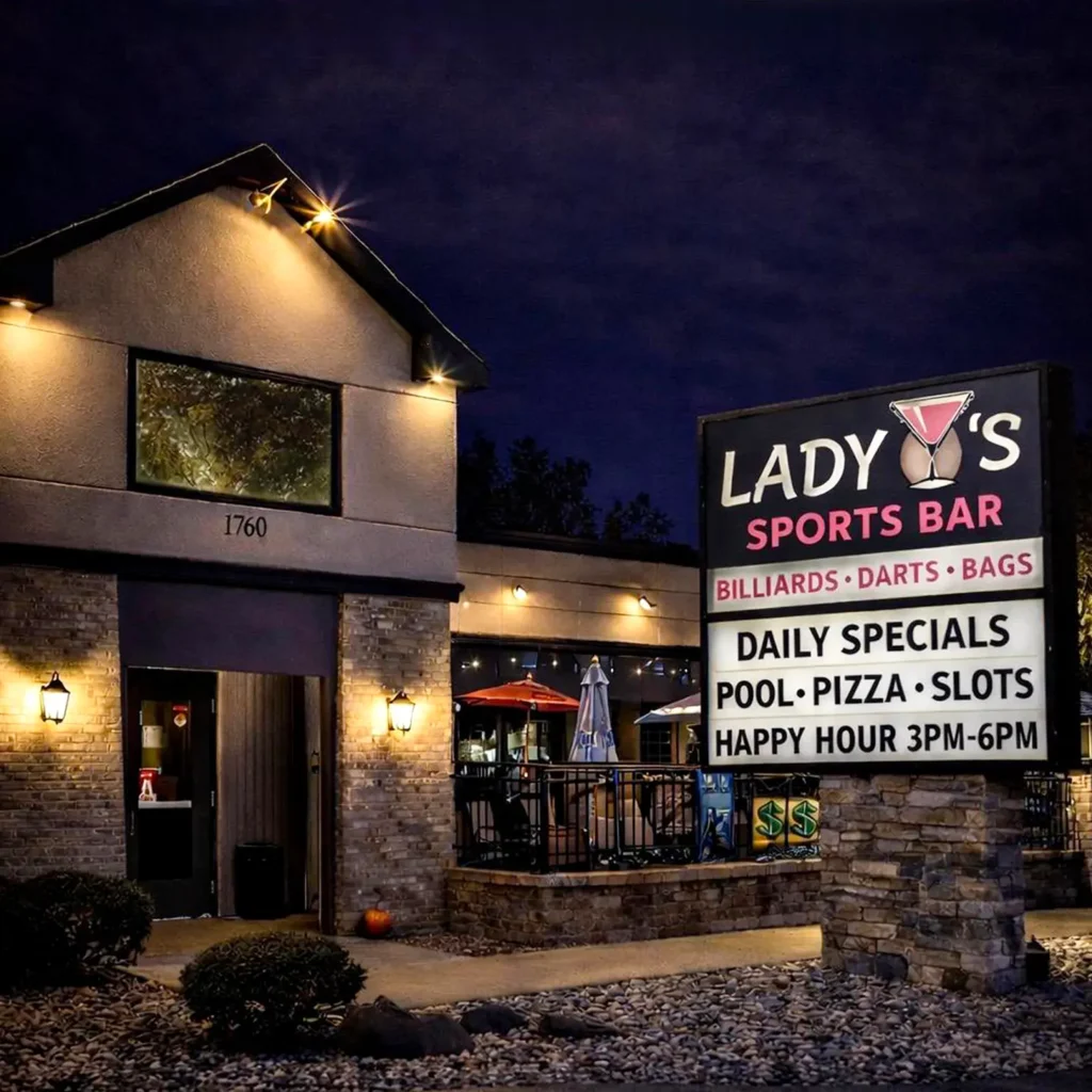 Fotografía nocturna de la fachada de "Lady's Sports Bar". El edificio es de ladrillo marrón y piedra, con una entrada de doble puerta de madera iluminada. A la derecha, destaca un letrero luminoso con un icono de una copa de cóctel que anuncia billar, dardos, pizza y "Happy Hour".